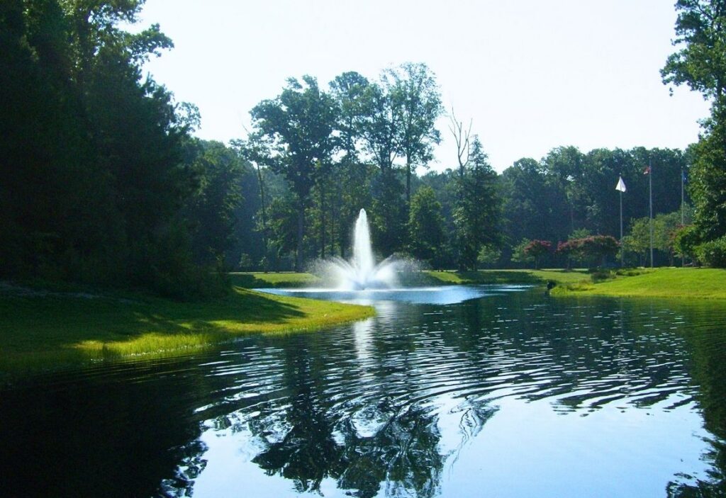 Pond with a spraying fountain, grassy shorelines, and mature trees reflected in the water