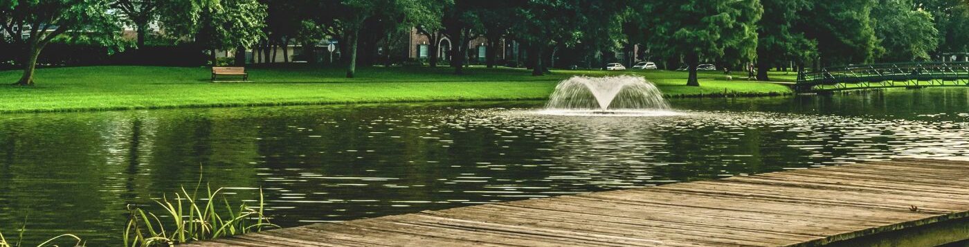 Pond with an umbrella-shaped fountain, a wooden boardwalk in the foreground, and a grassy shoreline lined with trees.