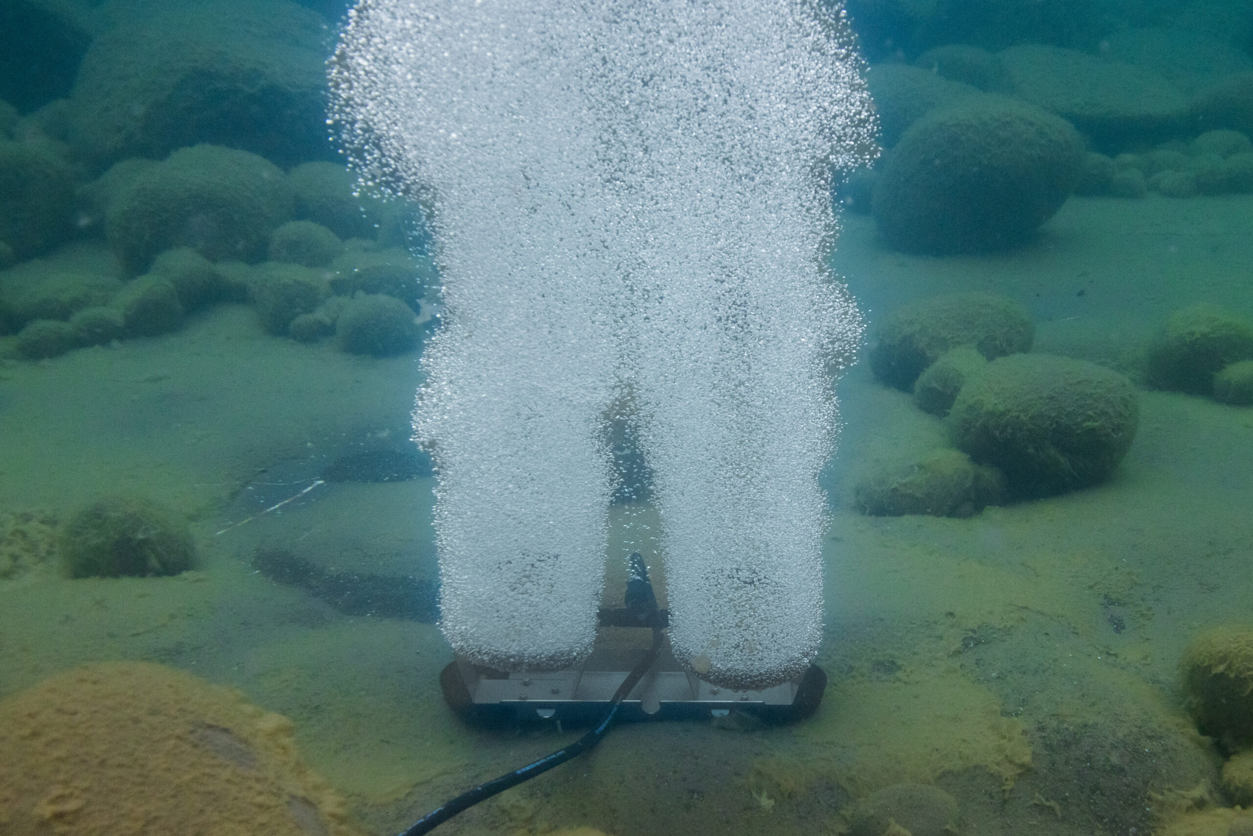 Submersed pond aerator releasing air bubbles from the bottom of a lake with algae-covered rocks nearby