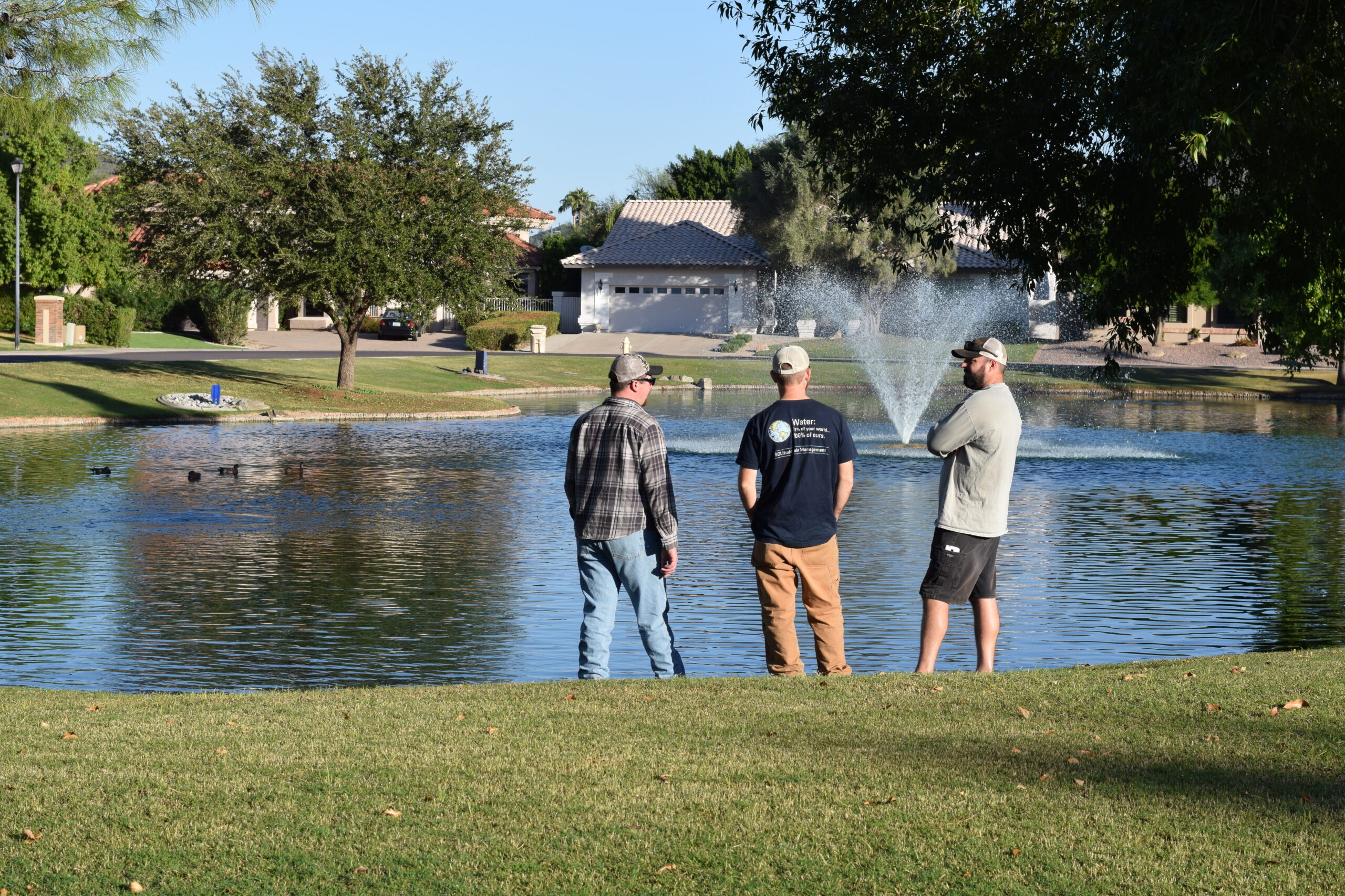 three men standing on edge of community pond with a fountain spraying in the background